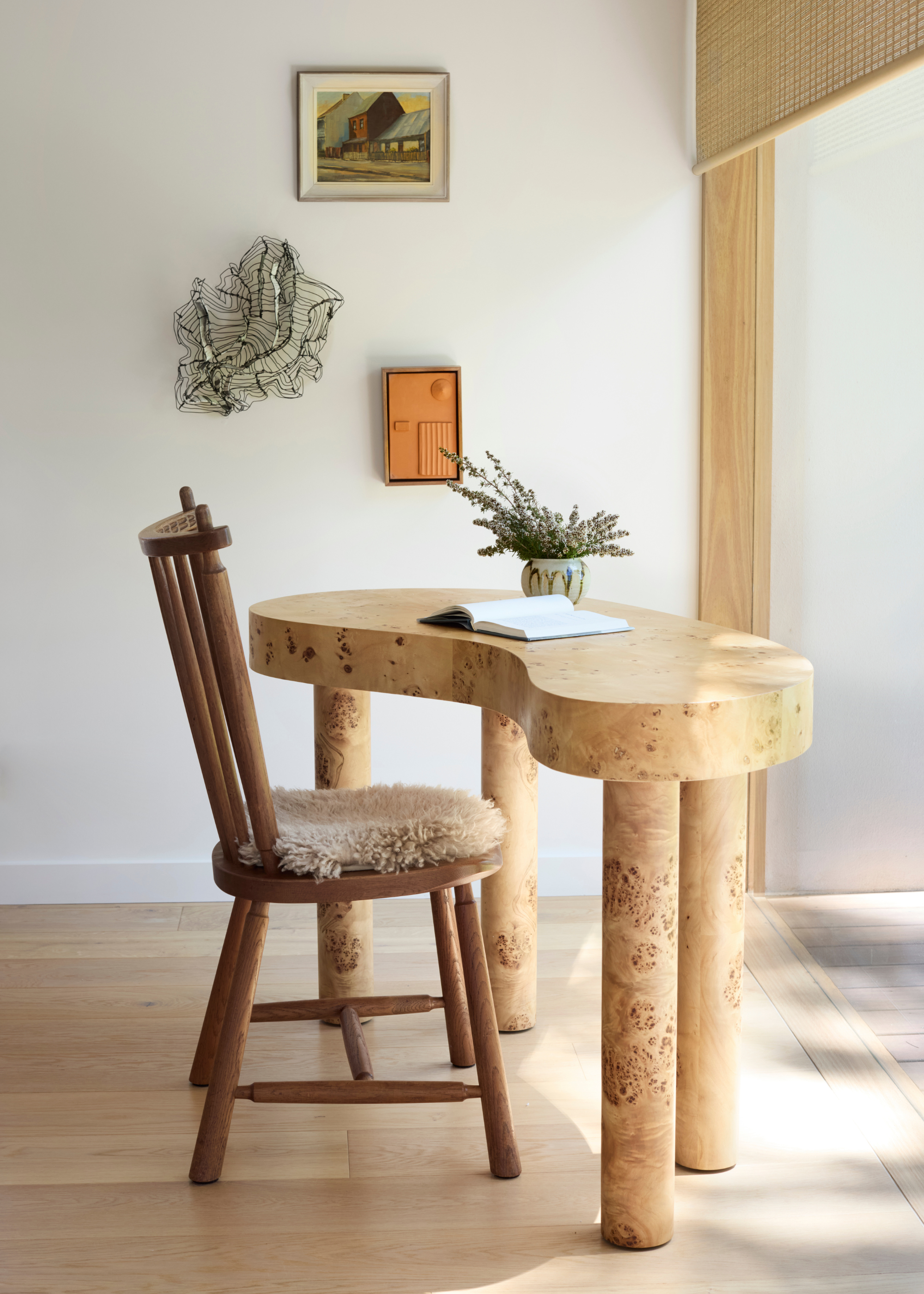 A study space with a curved burl wood table, a chair with a fur seat, a wall sculpture, and a pair of paintings