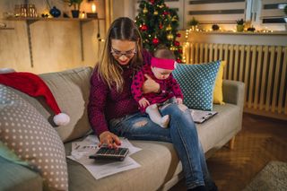 Mother and baby sat on sofa at Christmas with mother looking at bills and using a calculator