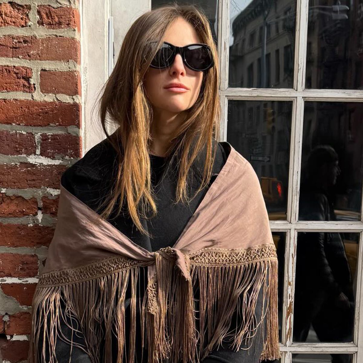 Hannah wears a black jacket with a brown fringe scarf over her shoulders.