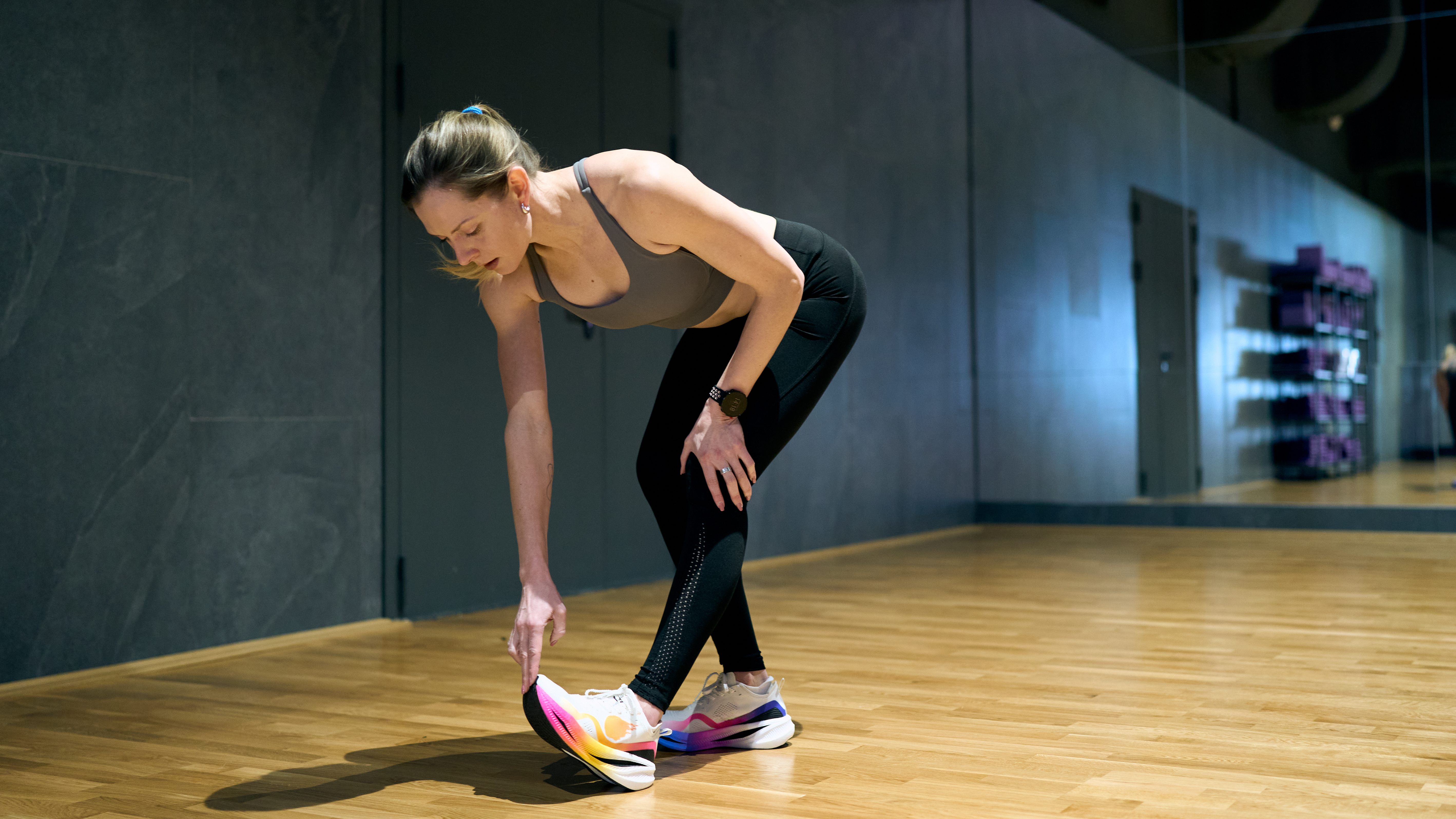 A woman in a sports bra, leggings and sneakers stretches in a gym. She is in a staggered stance, with her rear leg slightly bent and her left leg straight. She is hinged forward at the hip as she bends over to touch the lifted toes on her left foot. 