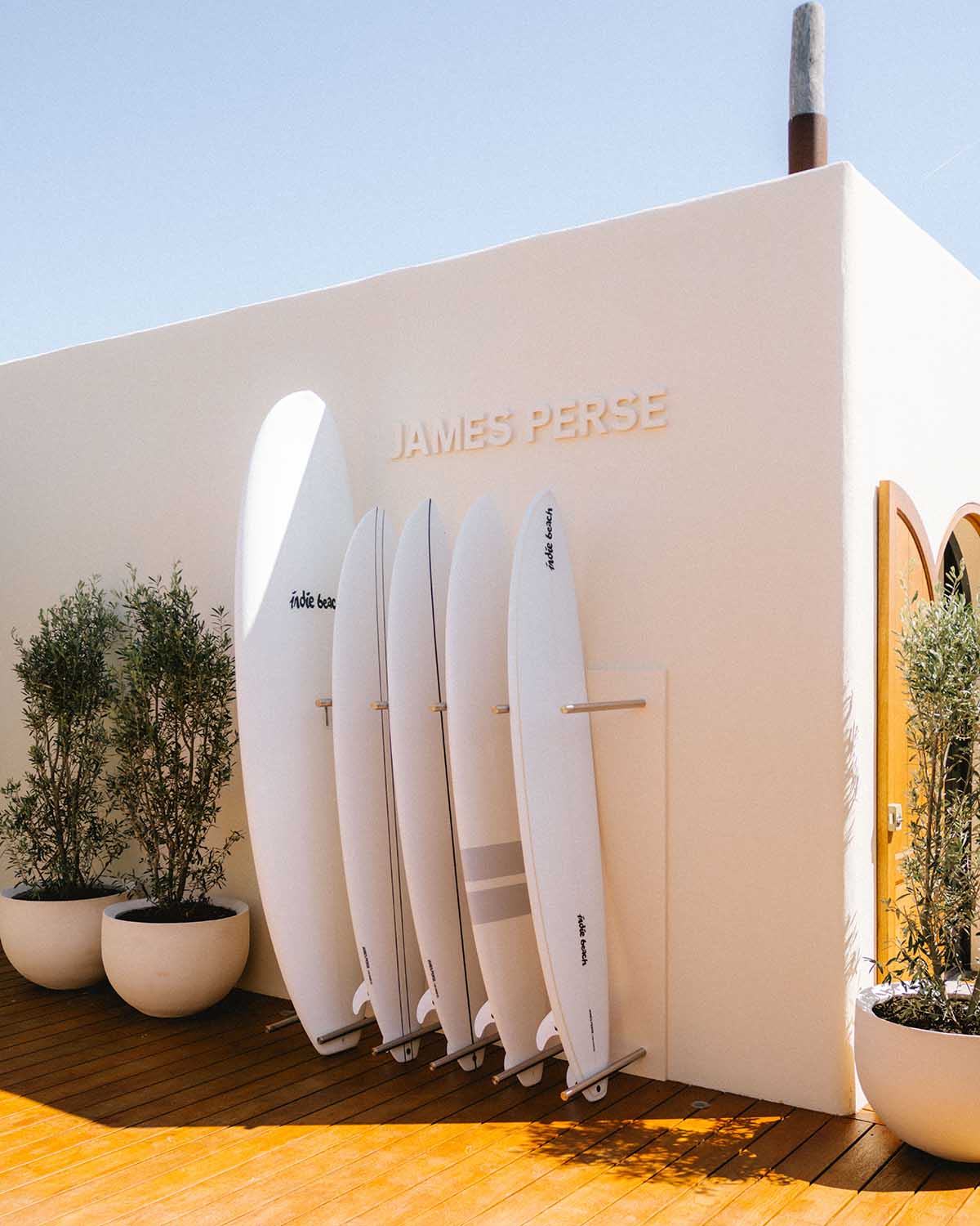 A series of white surfboards is displayed against a white wall on wooden flooring at a sun-filled beach club.