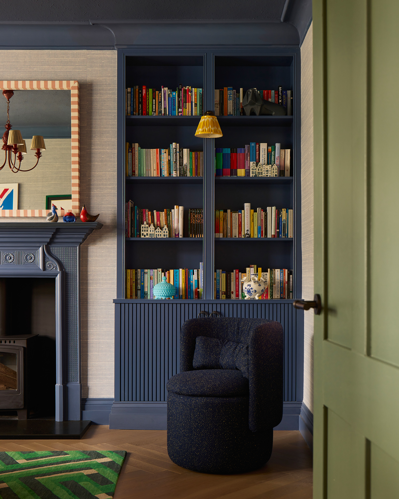 Image of the corner of a living room with a built-in bookcase that is painted dark blue and filled with colorful books. There is a navy accent chair in front of the bookcase and the trim in the room is also painted blue.