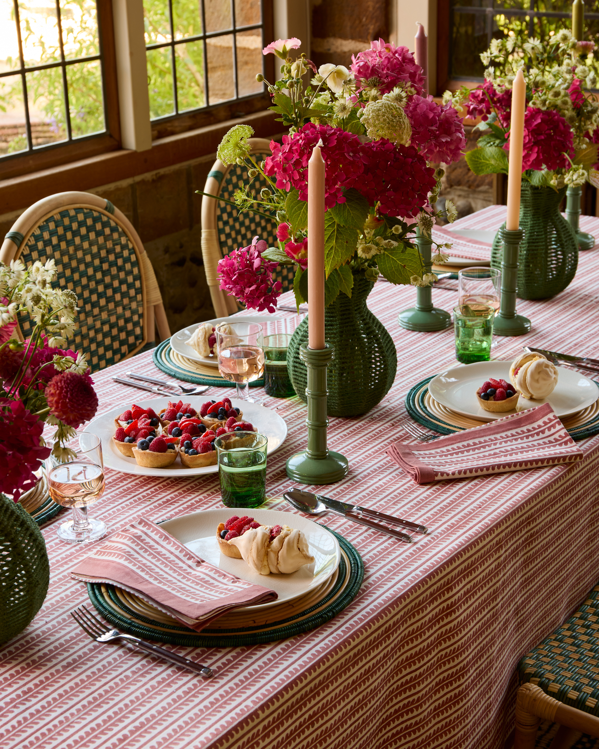 A table laid formally with a red patterned tablecloth, green candlesticks, and green rattan vases with pink hydrangeas in them