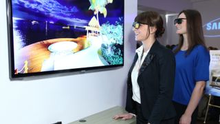 Two people wearing active 3D glasses watch a Samsung-branded TV. They are smiling and standing a few feet from the TV.