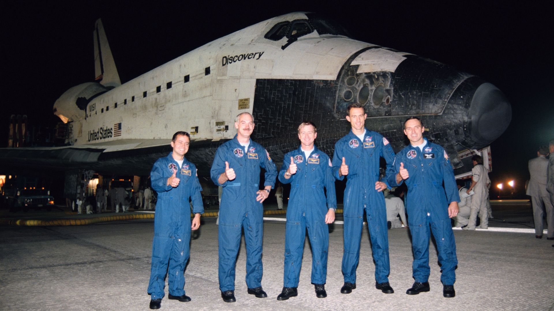 Uma tripulação de cinco astronautas em trajes de voo azuis levanta o polegar em frente a um ônibus espacial à noite
