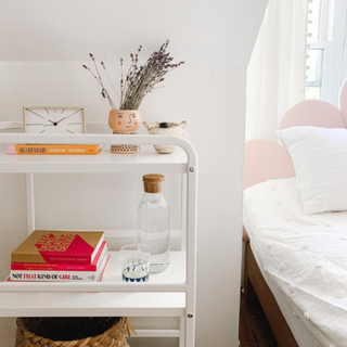 White bar cart utilized as bedside table, with books, alarm clock, water carafe, and decorative accessories.