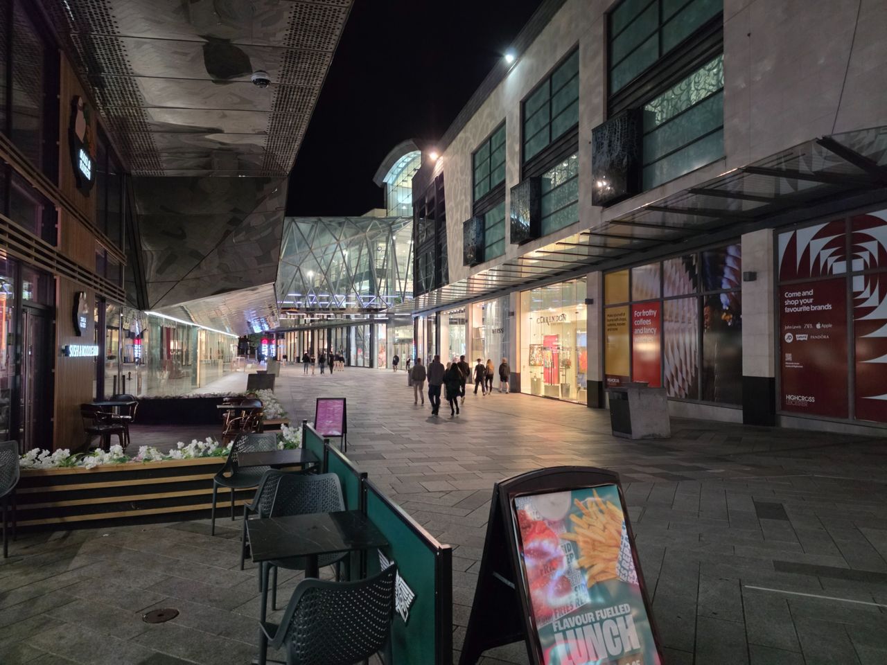 Nighttime shopping center walkway with brightly lit stores and people walking beneath modern glass architecture.