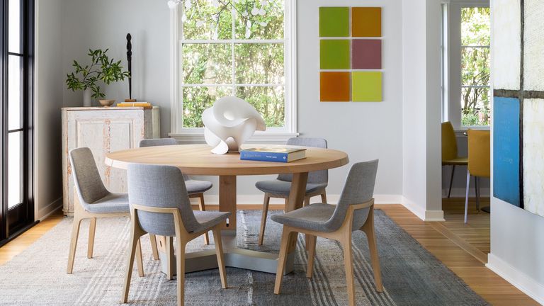 A dining room with a circular wooden table, grey upholstered chairs and multiple large abstract artworks on the walls