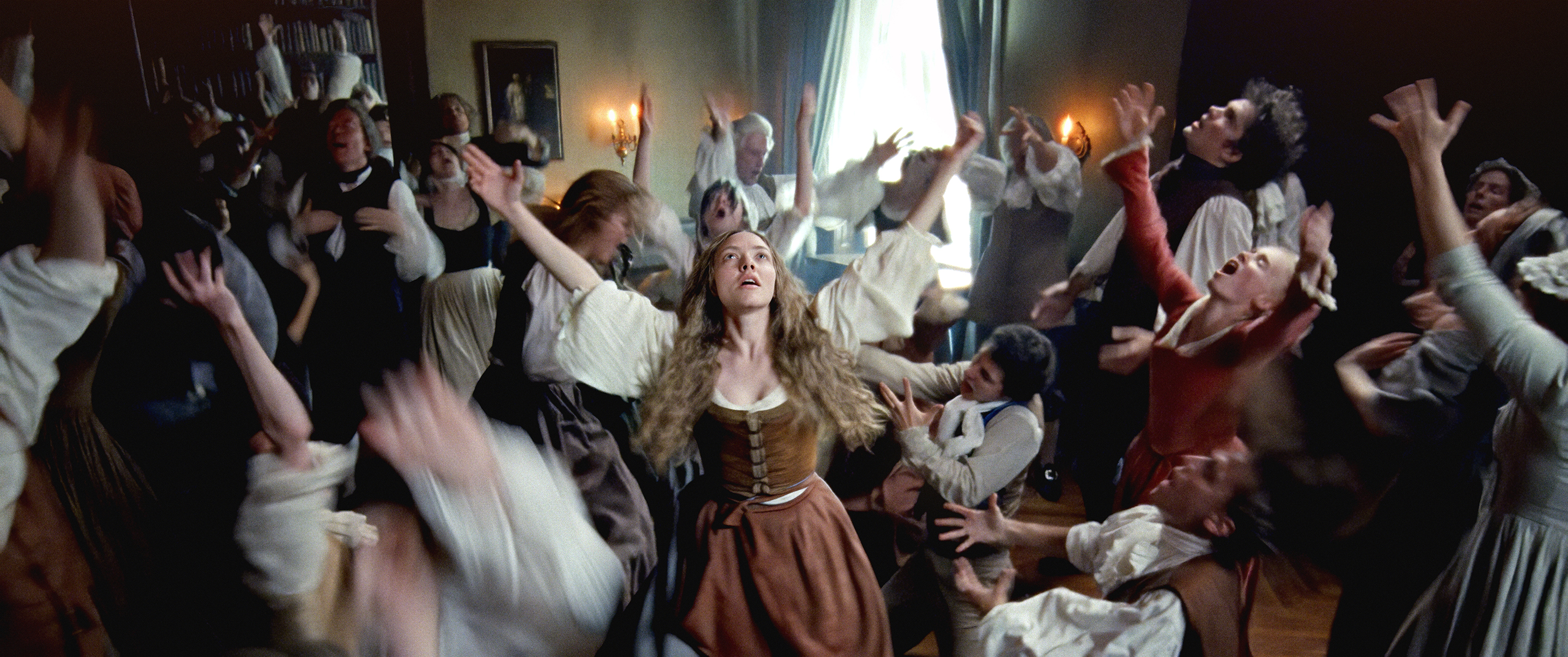 A group of people (center: Amanda Seyfried as Ann Lee) dressed in 18th-century clothing stand in a large room in an English manor, with a bookcase seen through a doorway. They all raise their heads and hands to the sky while dancing. Several people's limbs are blurred with movement, in a still from THE TESTAMENT OF ANN LEE.