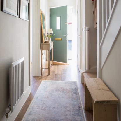 A narrow hallway with a faded-effect runner, a wooden bench and sage green front door