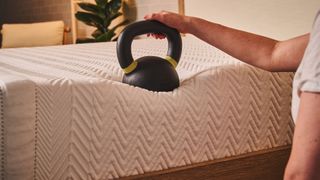 A 35lb kettlebell balances on the edge of the Leesa Legend Hybrid mattress, to test the edge support. It's sinking into the foams as a hand rests on the handle to keep the weight in place