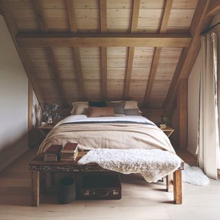 A bedroom with a slanted ceiling with beams and a bed decorated with neutral-coloured cushions and a throw