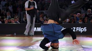 a b-boy in a black and blue jacket spins on his head in front of a crowd