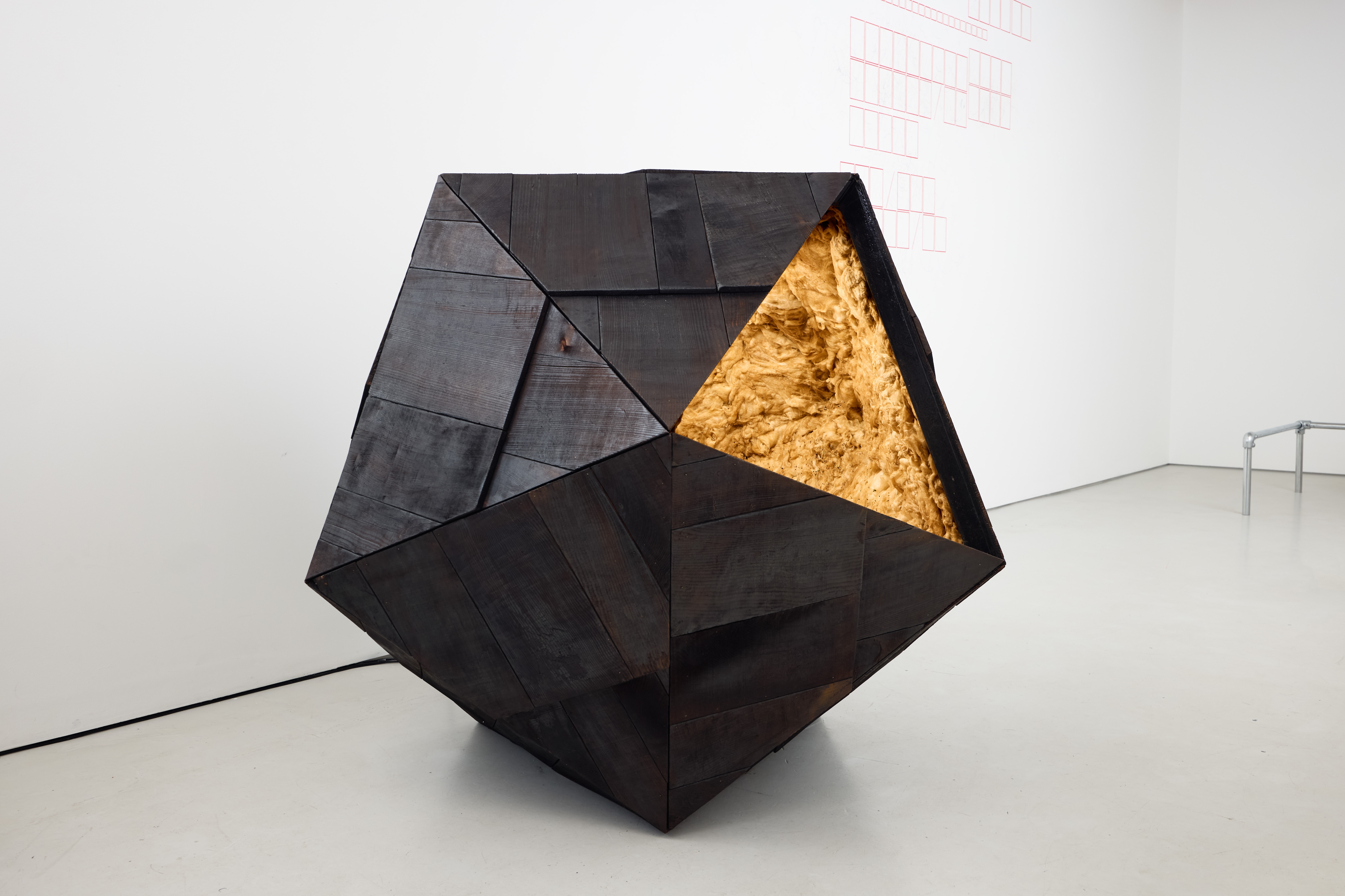 A prism-shaped sculpture made of wood and golden-looking fibers sits in an empty gallery room.