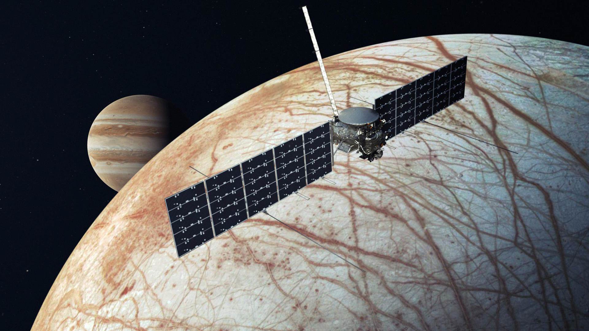 An image of the Europa clipper spacecraft. It has a small body with wide wings. Beneath it, Europa is tan and brown with a vein-like pattern.