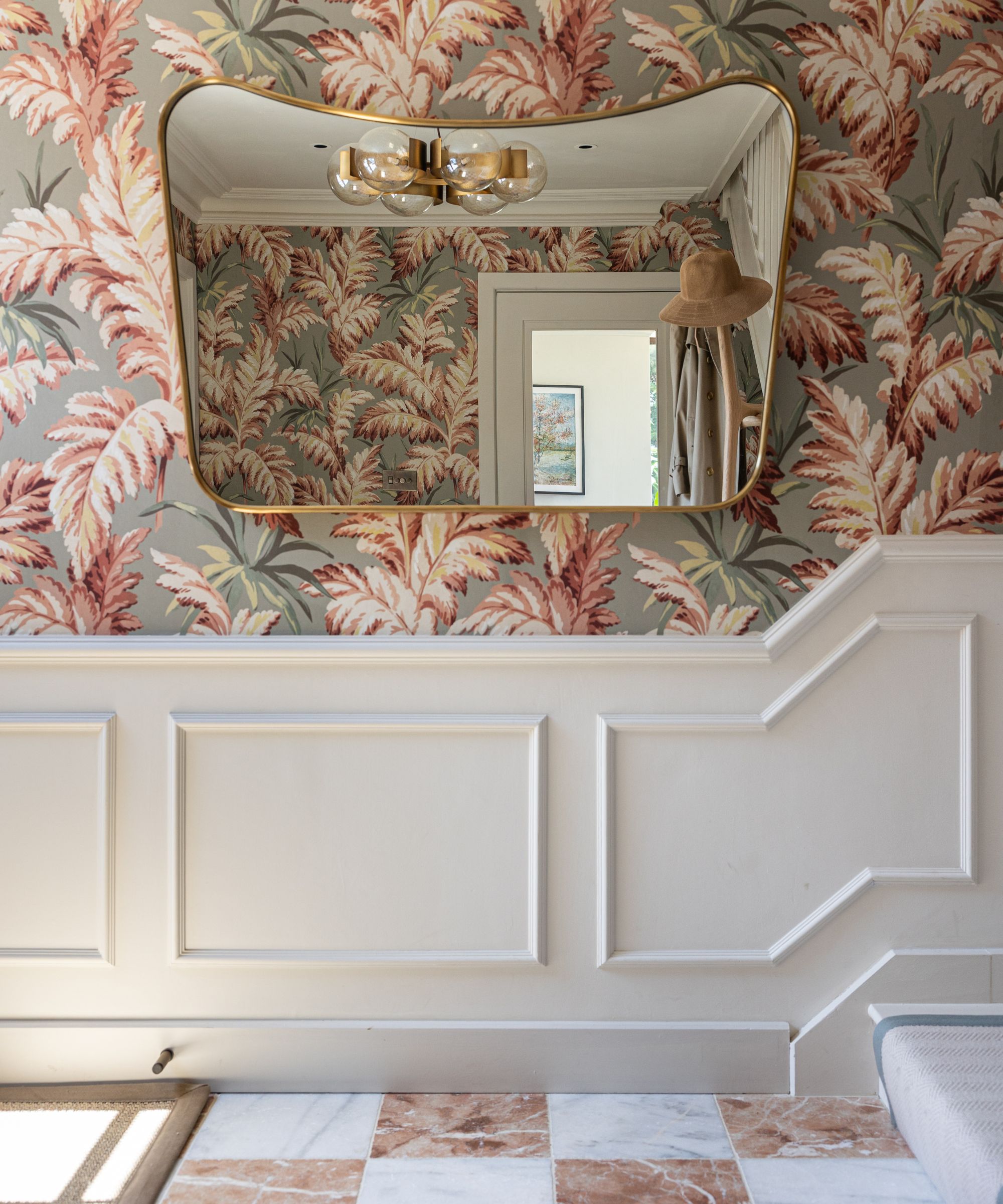 Victorian-style hallway with bold botanical wallpaper, ornate white panelling and marble checkerboard flooring, reflected in a curved brass-framed mirror.