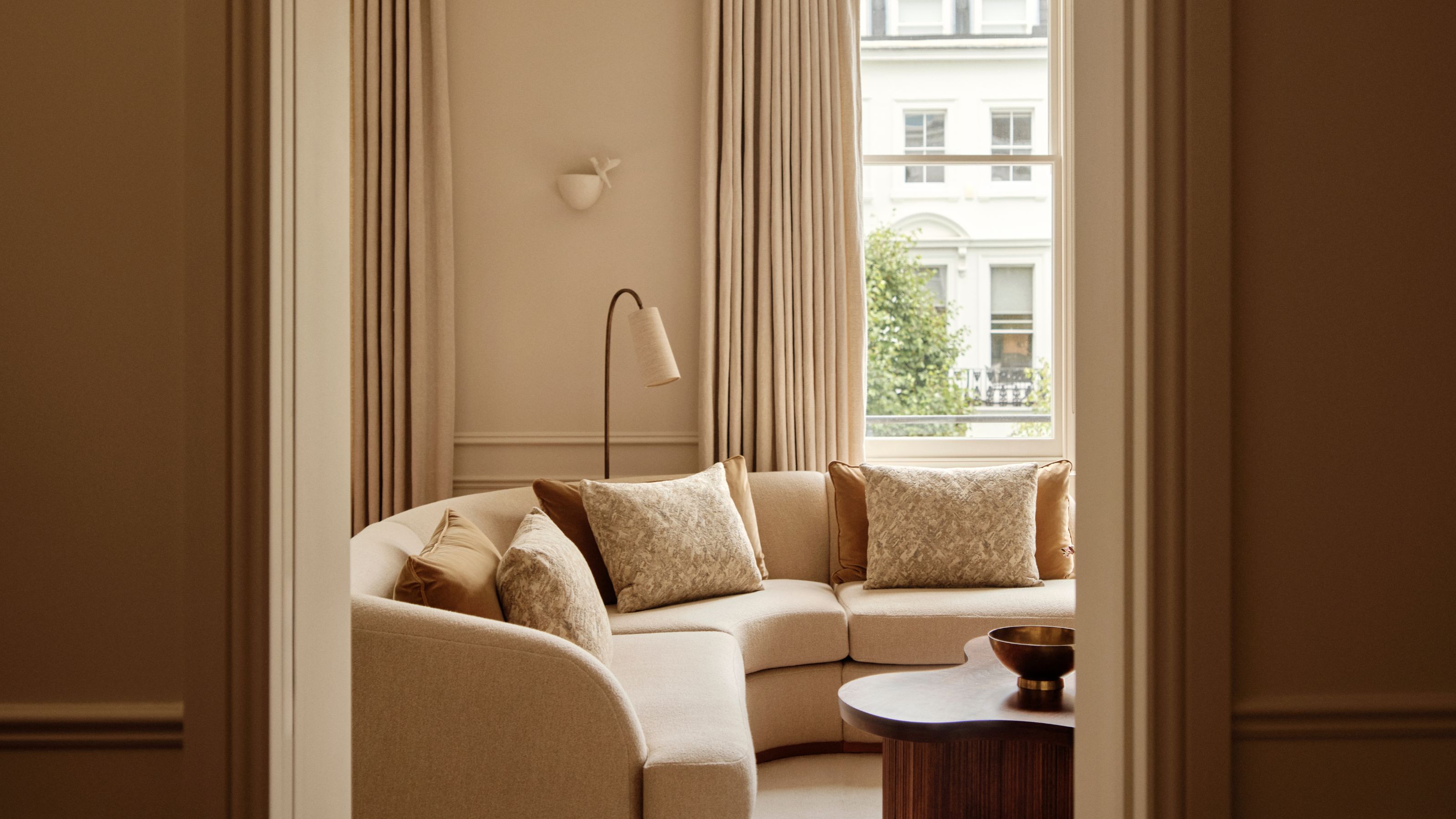 A doorway leading into a modern, neutral living room in a period home with soft neutral walls and curtains, an arched window, a large beige corner sofa, a dark wood sculptural coffee table and a cream rug.