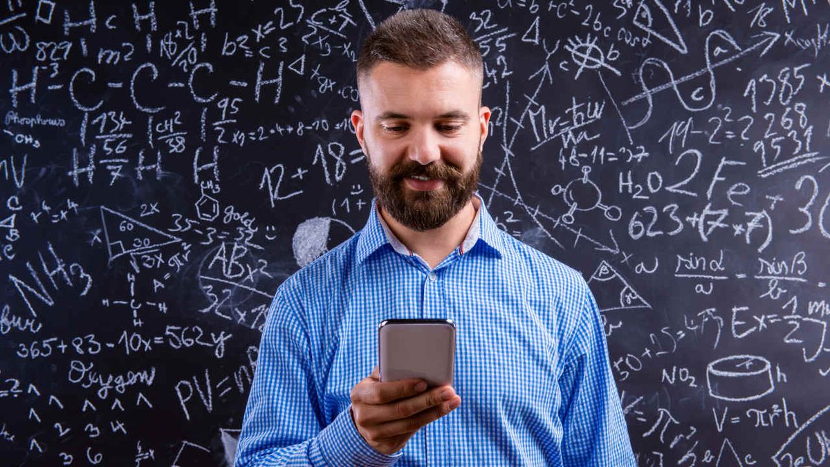 Smiling, bearded teacher looks at cell phone while standing in front of blackboard covered with chemistry equations