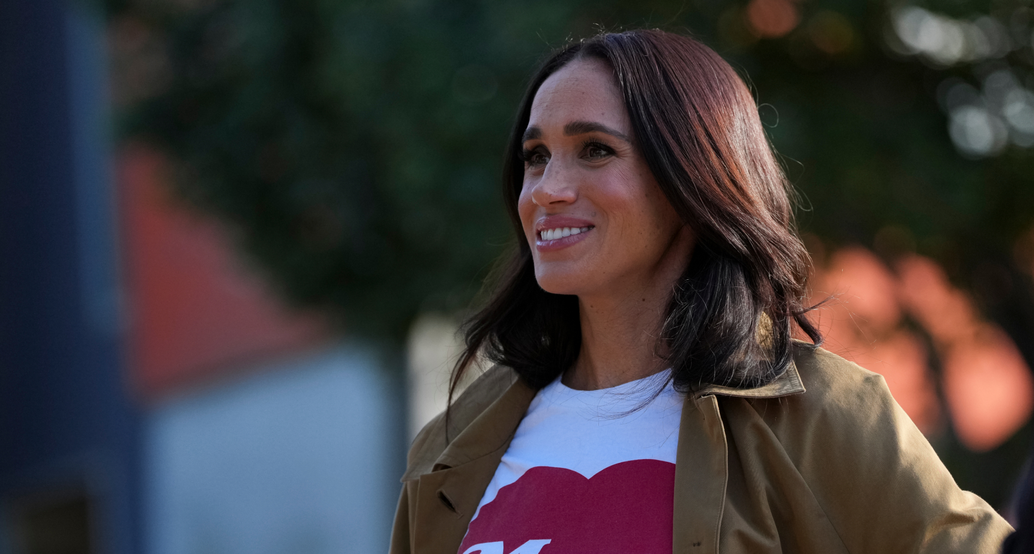 Meghan Markle smiling wearing a white and red t-shirt and a trench coat