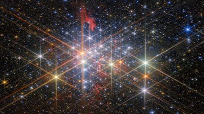 A dense cluster of bright stars, each with six large and two small diffraction spikes, due to the telescope&rsquo;s optics. They have a variety of sizes depending on their brightness and distance from us in the cluster, and different colors reflecting different types of star. Patches of billowing red gas can be seen in and around the cluster, lit up by the stars. Small stars in the cluster blend into a background of distant stars and galaxies on black.