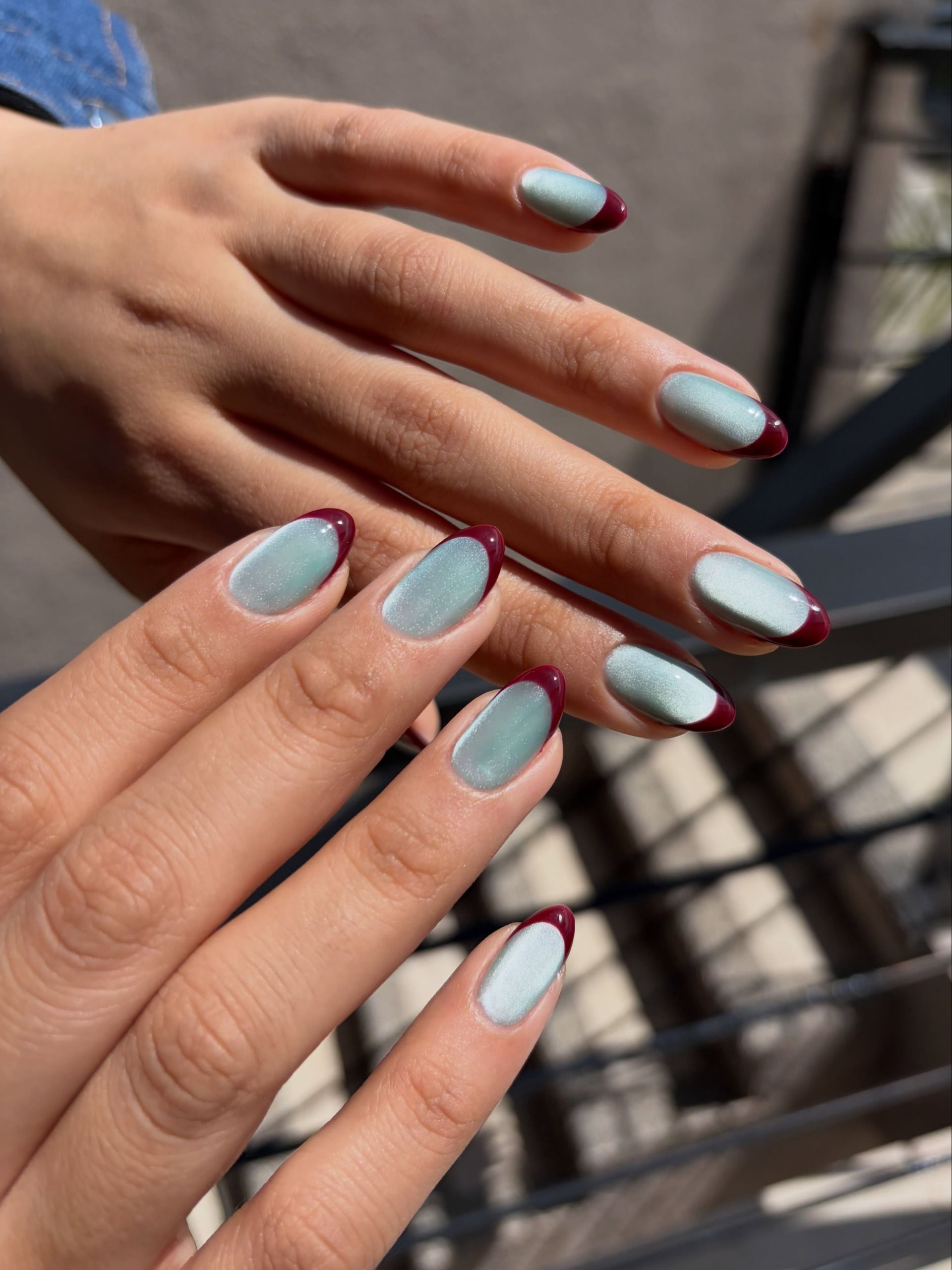 Baby blue almond nails with thin burgundy French tips, hands resting in the sun