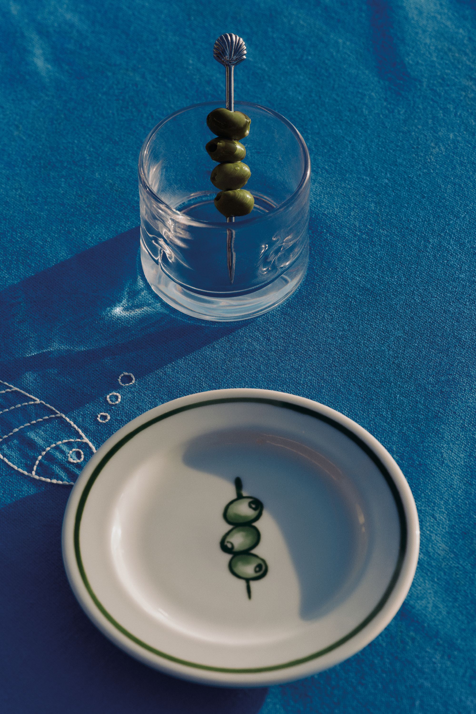 Image of a plate with a drawing of olives in the center of it and a cocktail glass with olives on a pick. The dinnerware is sitting on a deep blue tablecloth.