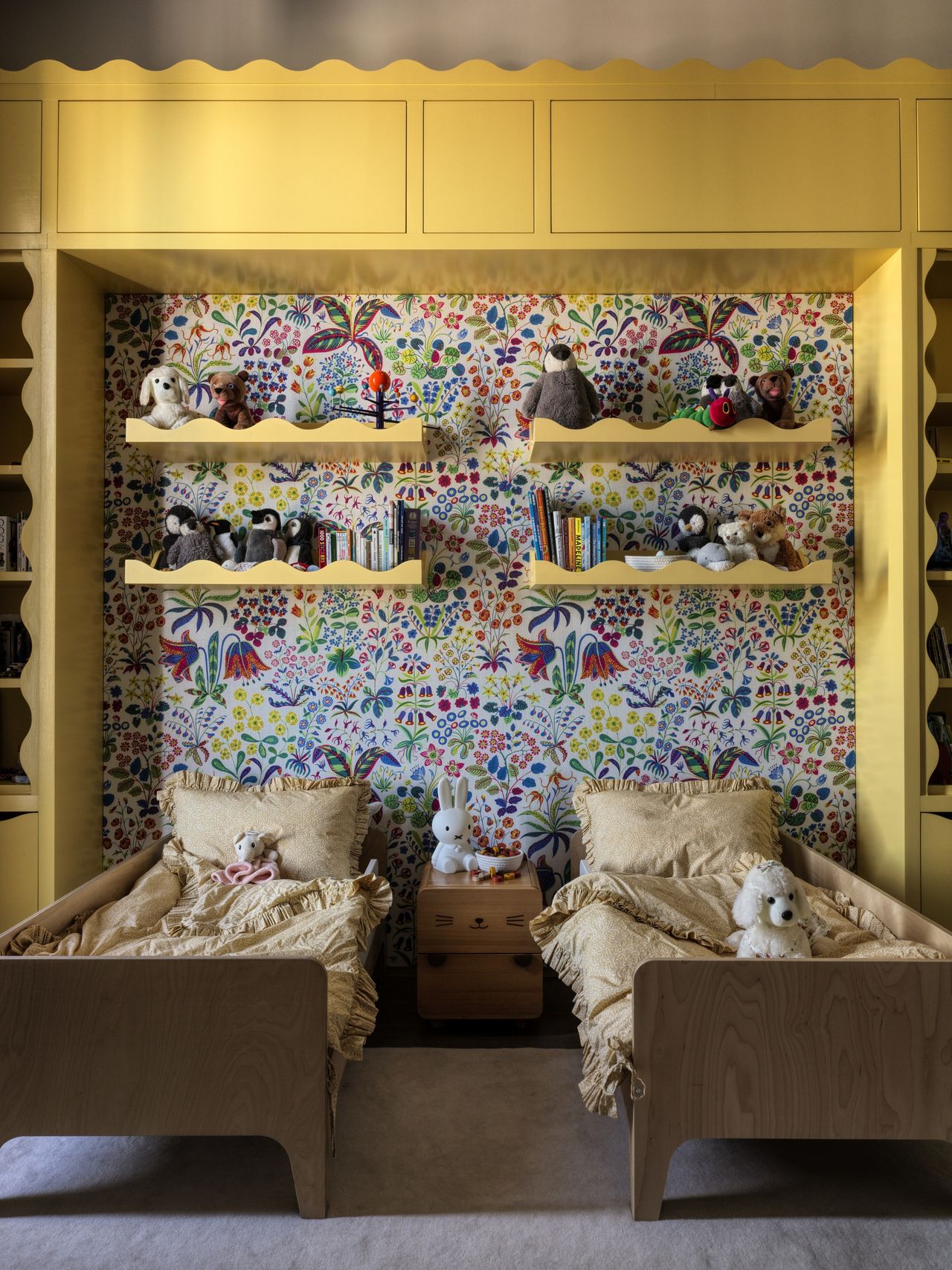 kids bedroom with yellow built-in cabinetry with scalloped edges, floral wallpaper, floating scallop shelves with toys and books, and two timber twin beds with yellow sheets