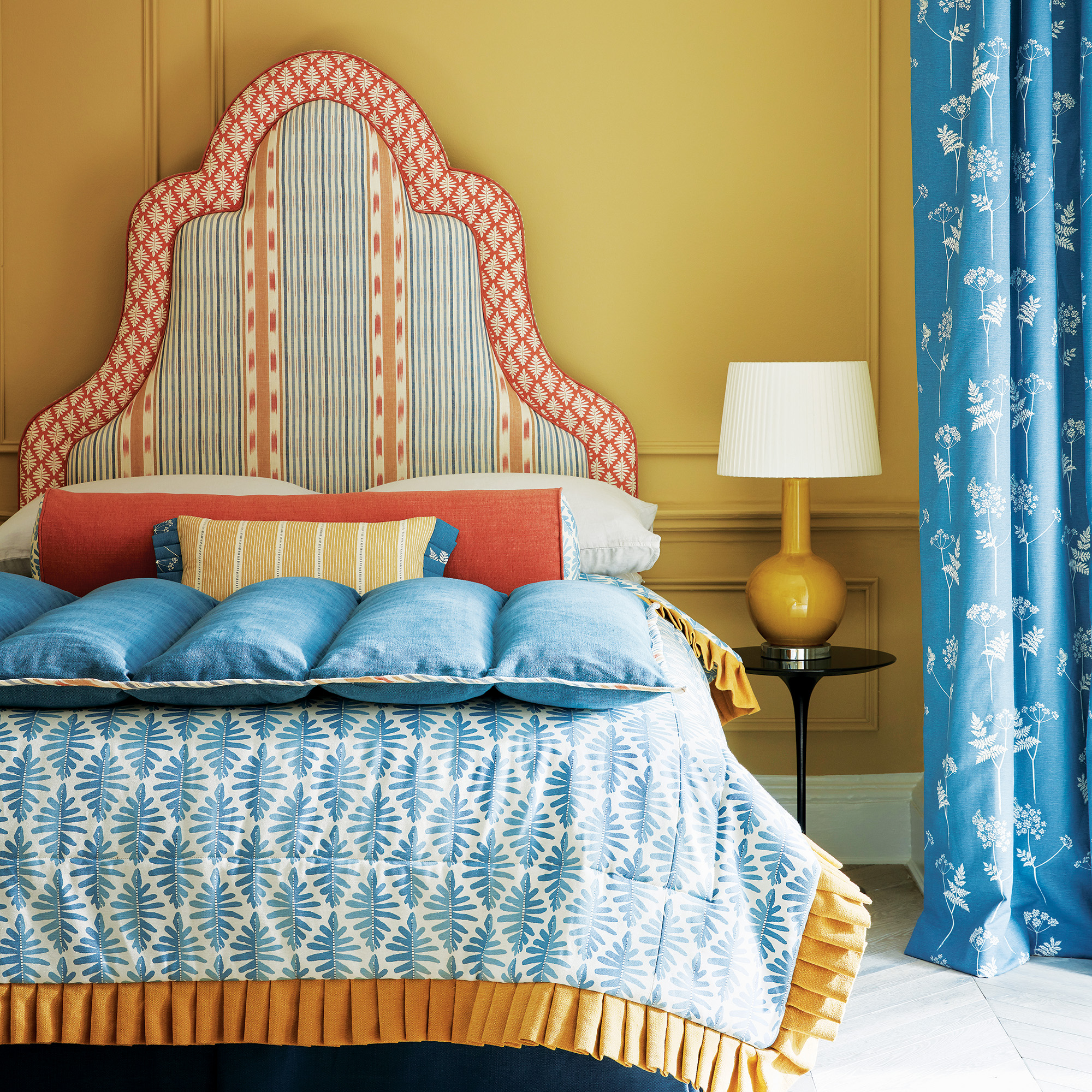 bedroom with yellow panelled walls and blue curtains and a statment bed with sculputral patterned headbaord and blue and orange themed bedlinens and cushions vanessa arbuthnott