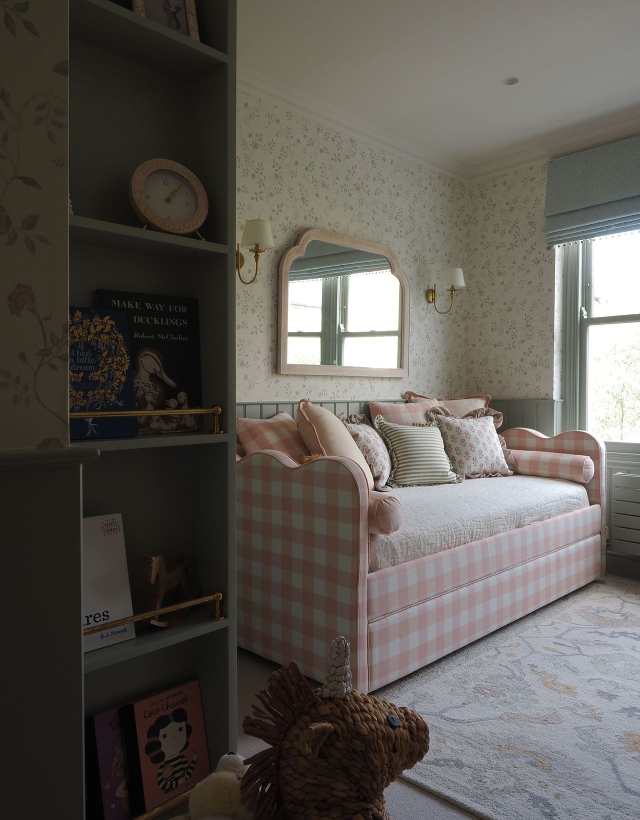 A more traditional-style bedroom with a pink and white checked daybed with lots of cushions on top and alcove shelving in the foreground with books and decorative objects next to a toy unicorn