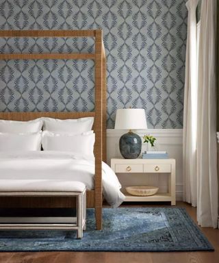 White bedding on a wood frame four-poster; a lamp on a white nightstand to the right of the bed; a blue rug on the floor; all against white and blue wallpaper.