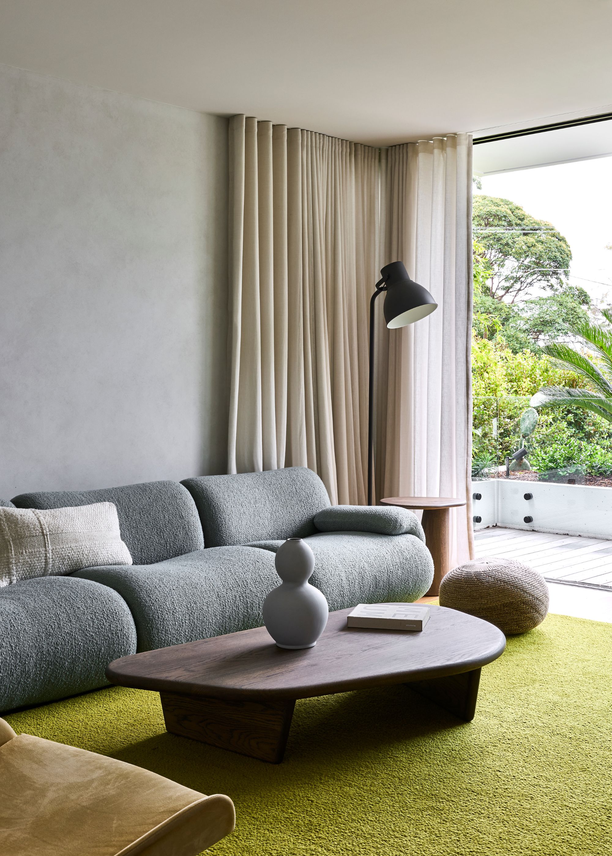 A living room with white curtains, a black floor lamp, a green area rug, a blue couch, a wood coffee table, a vase, and a white pillow