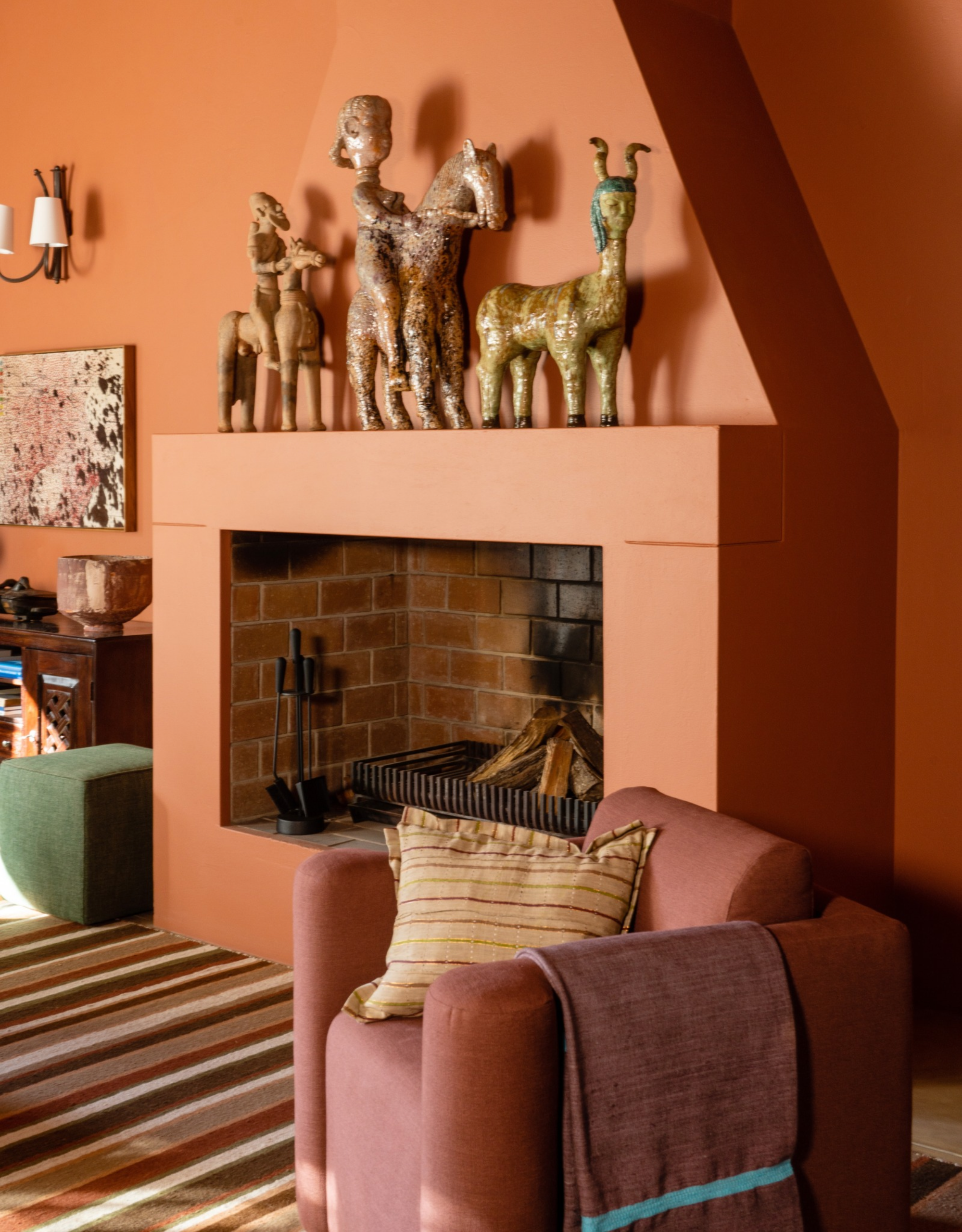 A terracotta-colored room with a fireplace, a striped rug, and a plum armchair. Also pictured is a row of ceramic arts over the mantel