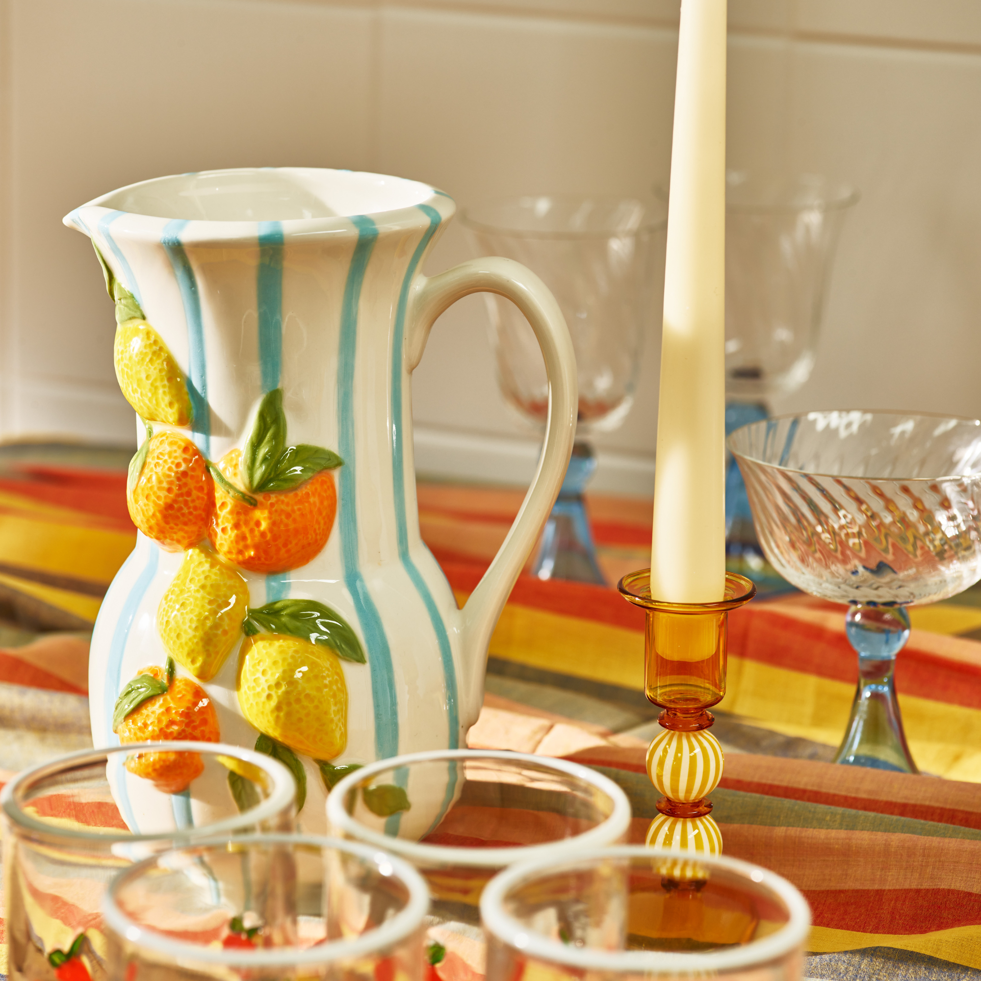 Blue and white striped jug with 3D fruit attached. There is an orange and white striped glass candlestick next to it and cokcktail glasses in the foreground and background.