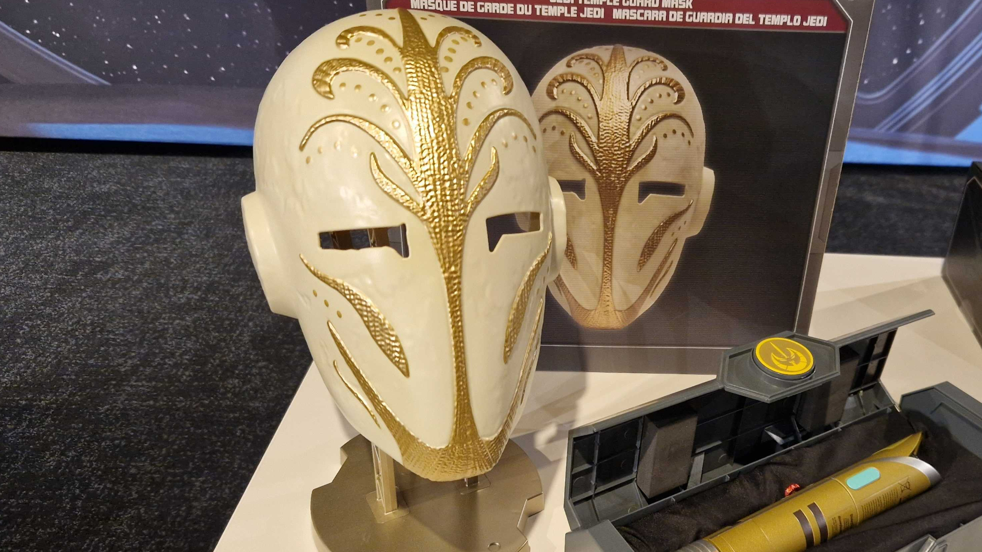 Star Wars Jedi Temple Guard Mask on a stand, in front of a box and a lightsaber case