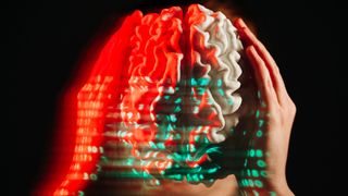 A person holds a white model of a brain with their hands on either side while lines of green and red binary numbers are projected on top
