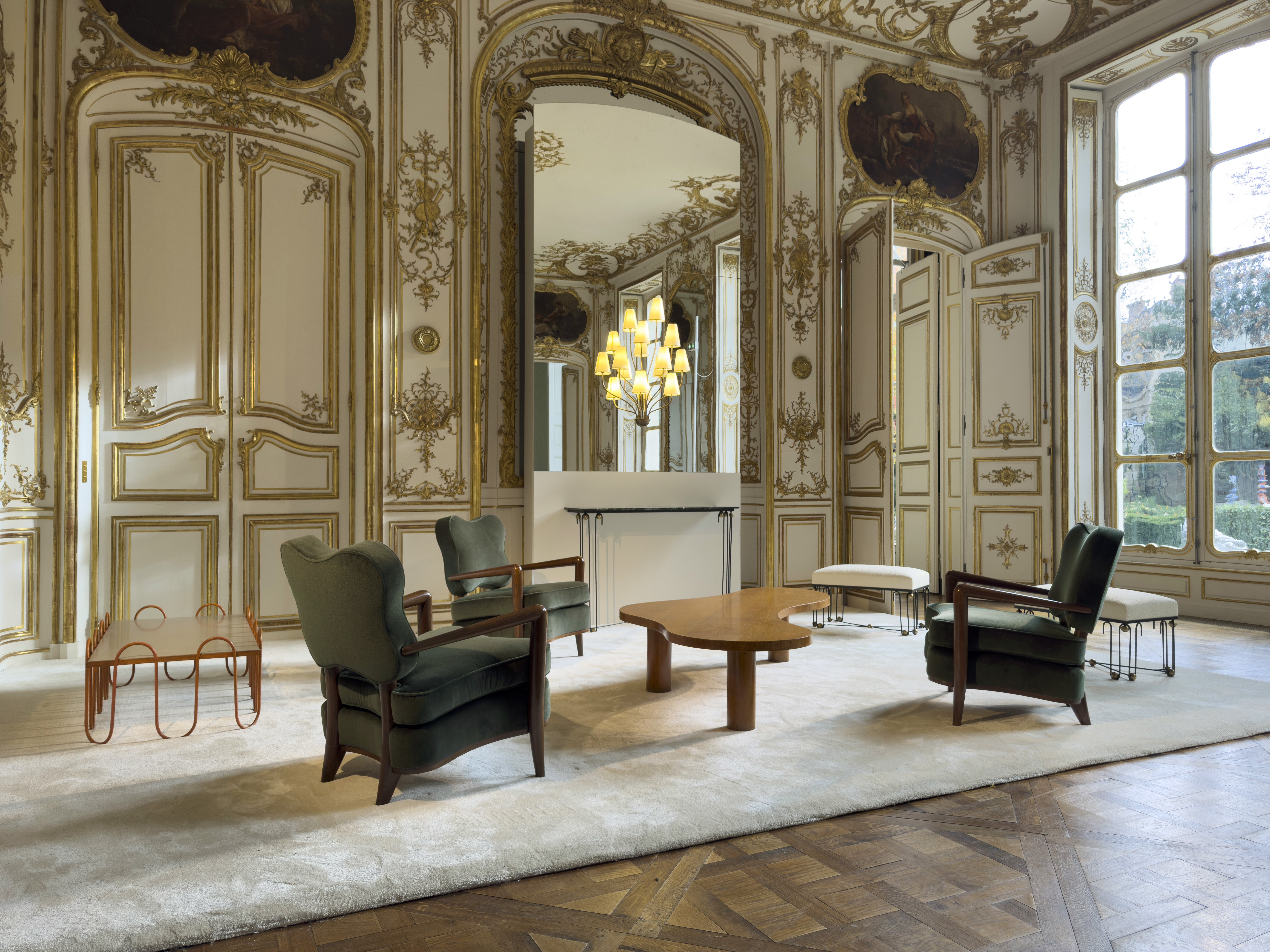 A stucco and gold leaf-decorated, historical building houses a series of mid-century furniture items, including wavy wooden coffee tables, wood and textiles armchairs, and upholstered ottomas, in a naturally lit environment.