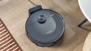 a rechargeable robot vacuum and mop in black by ultenic is photographed on a hardwood floor with a blue wall and a burnt orange terracotta and white striped rug