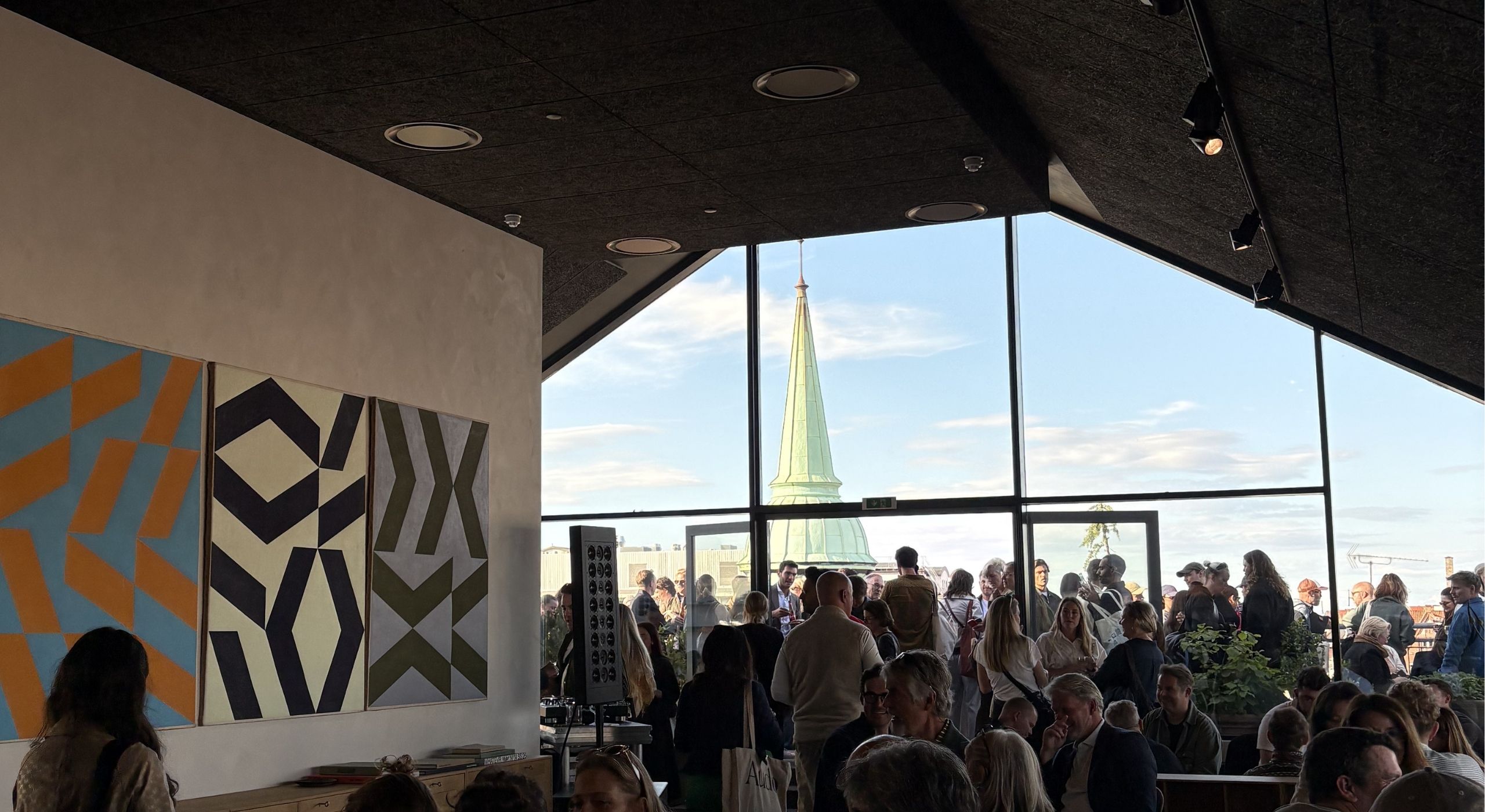 A rooftop bar with a sun-lit view over a historical city, seen from inside and filled with people mingling and abstract artworks.