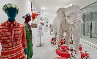 Mannequins displaying African clothing and handbags next to a large sculpted elephant standing above a large red snake with shoes displayed on it.