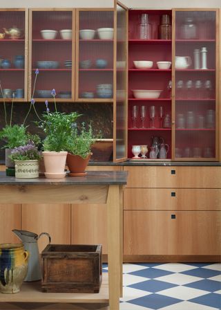 White and blue kitchen flooring contrasts beautifully with fluted glass kitchen cabinets that have been painted red inside. Opposite the cabinets is a light oak kitchen island with a stone countertop.