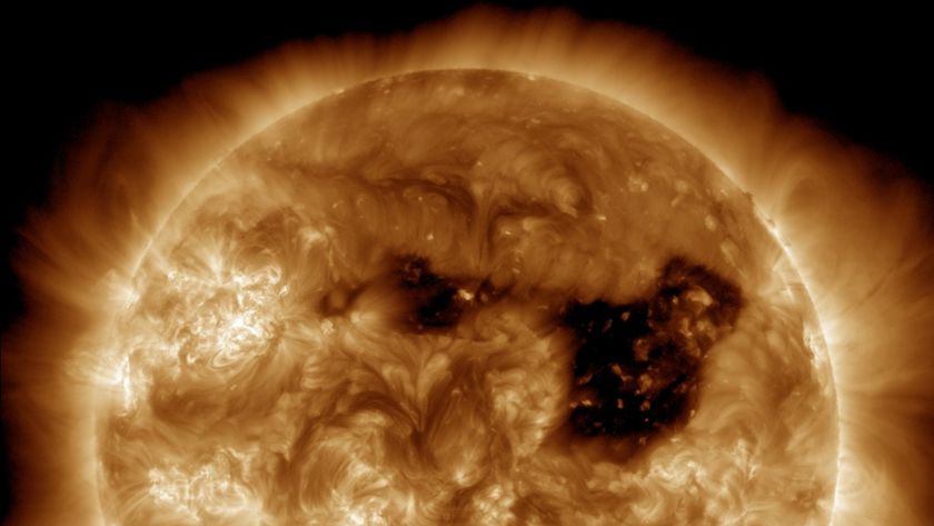 A high-resolution image of the sun taken in extreme ultraviolet light, showing a massive dark coronal hole on its surface. The coronal hole appears as a large black region where the sun’s magnetic field has opened up, allowing solar wind to escape into space. Bright swirling plasma structures surround the dark area, and the sun&#039;s outer atmosphere, or corona, glows in shades of gold and orange.