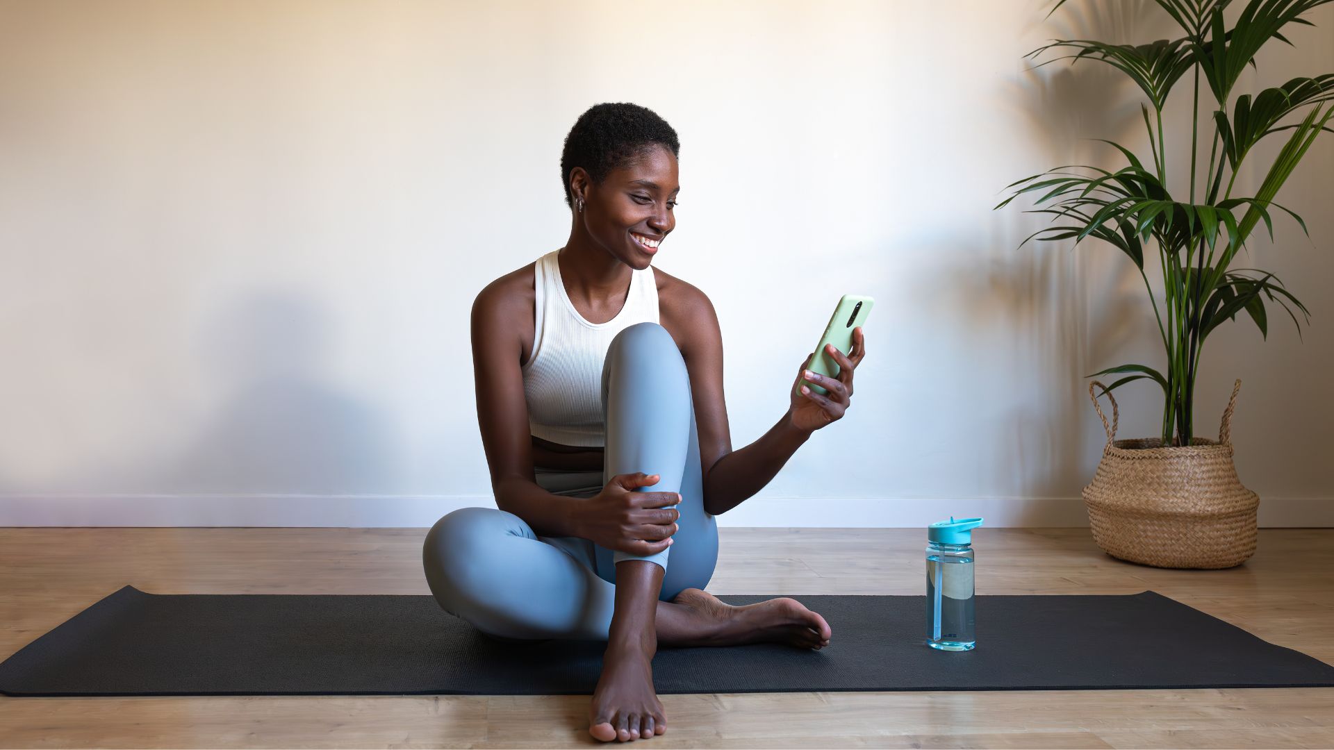 Woman looking at phone and smiling, sitting on yoga mat next to plant at home in activewear, setting up for the fire hydrant exercise