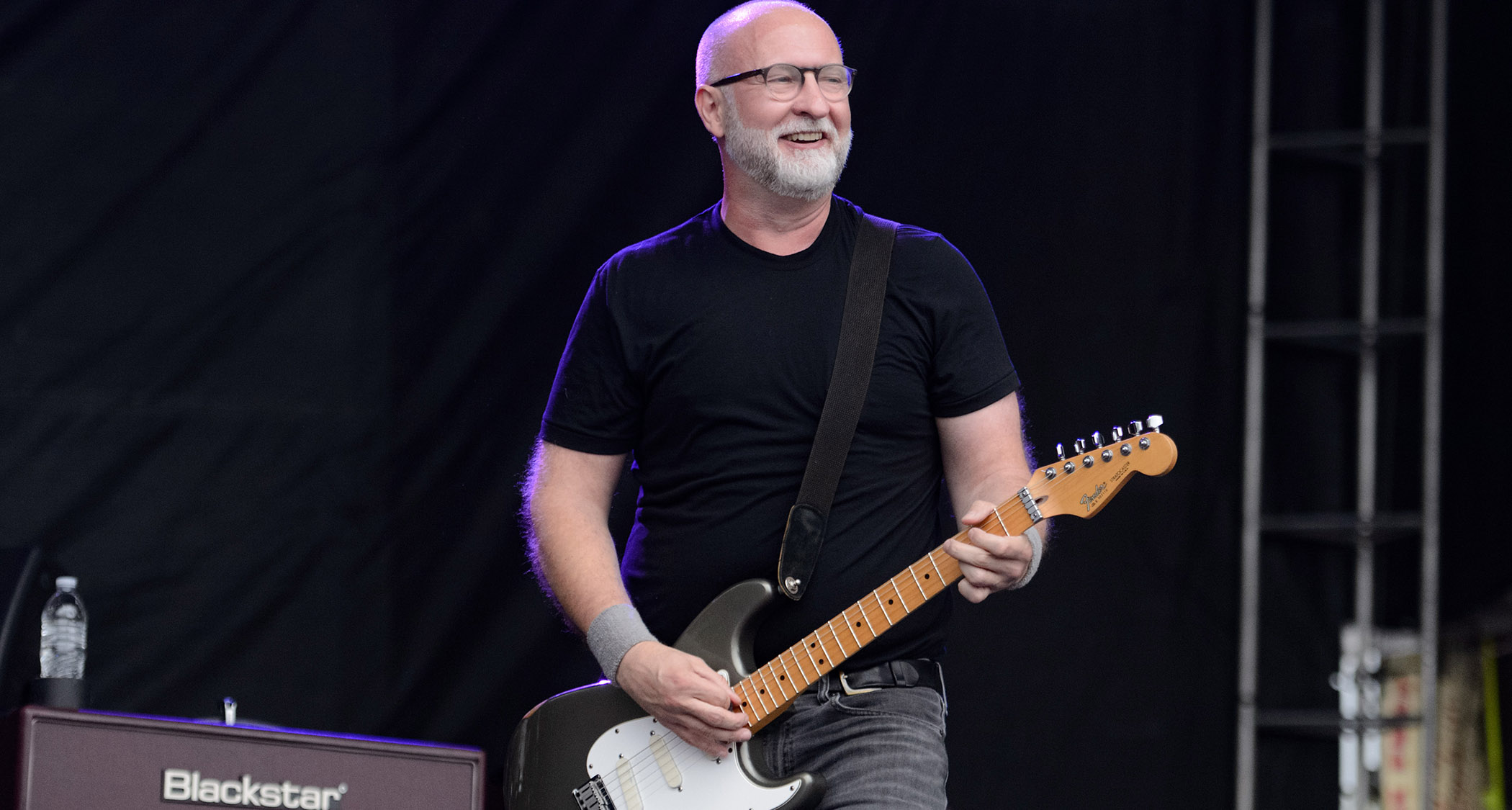 Bob Mould plays a gray Strat onstage at an outdoor gig. He wears a black T-shirt and glasses, and there is a Blackstar head in the background
