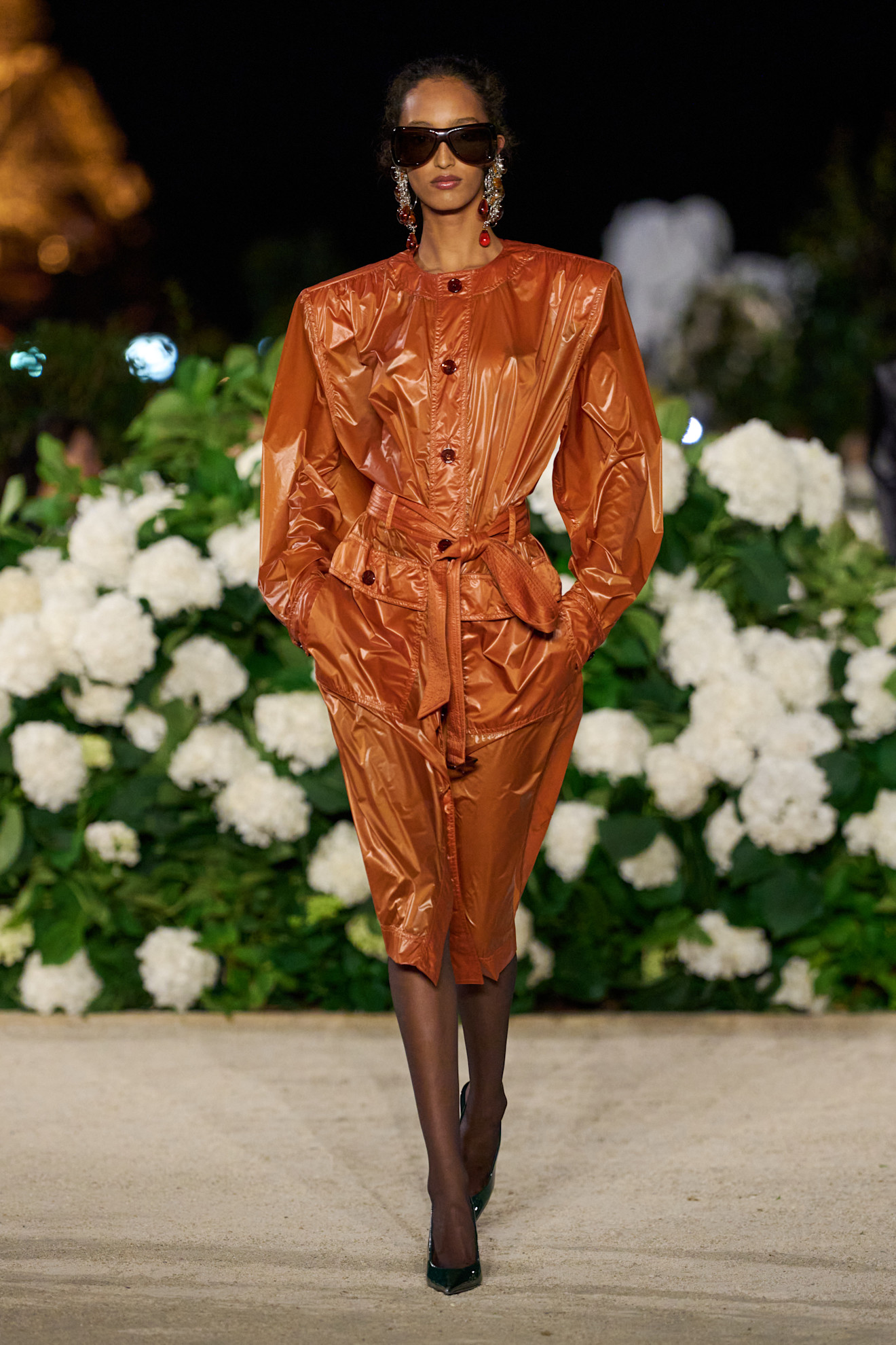 A Saint Laurent model wearing a dark orange trench coat styled as a dress, black sheer tights, black pointed pumps, black aviator sunglasses, and chain earrings with orange and red resin charms.