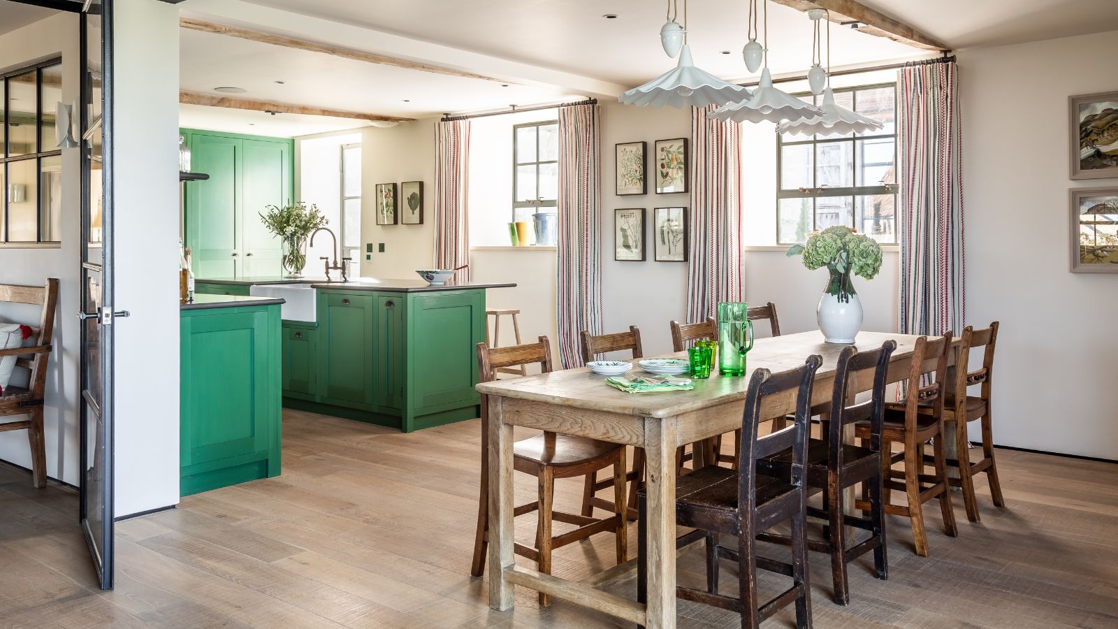 Large kitchen and dining area with eight-seated wooden table and wooden chairs, hanging white lampshades, exposed wooden ceiling beams, green kitchen cabinets and a wooden floor. Small windows brightly light up the space.