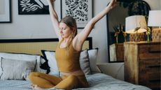 white woman wearing mustard top leggings stretching in bed bedroom background