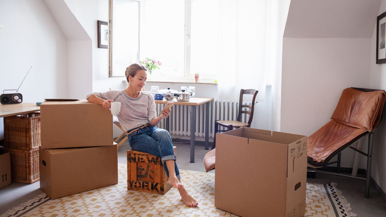 An older woman looks at her phone while sitting among moving boxes in her living room.