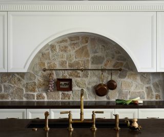 An exposed stone brick kitchen backsplash with a brass pot rail with pans and an inherited framed art piece hung from it.