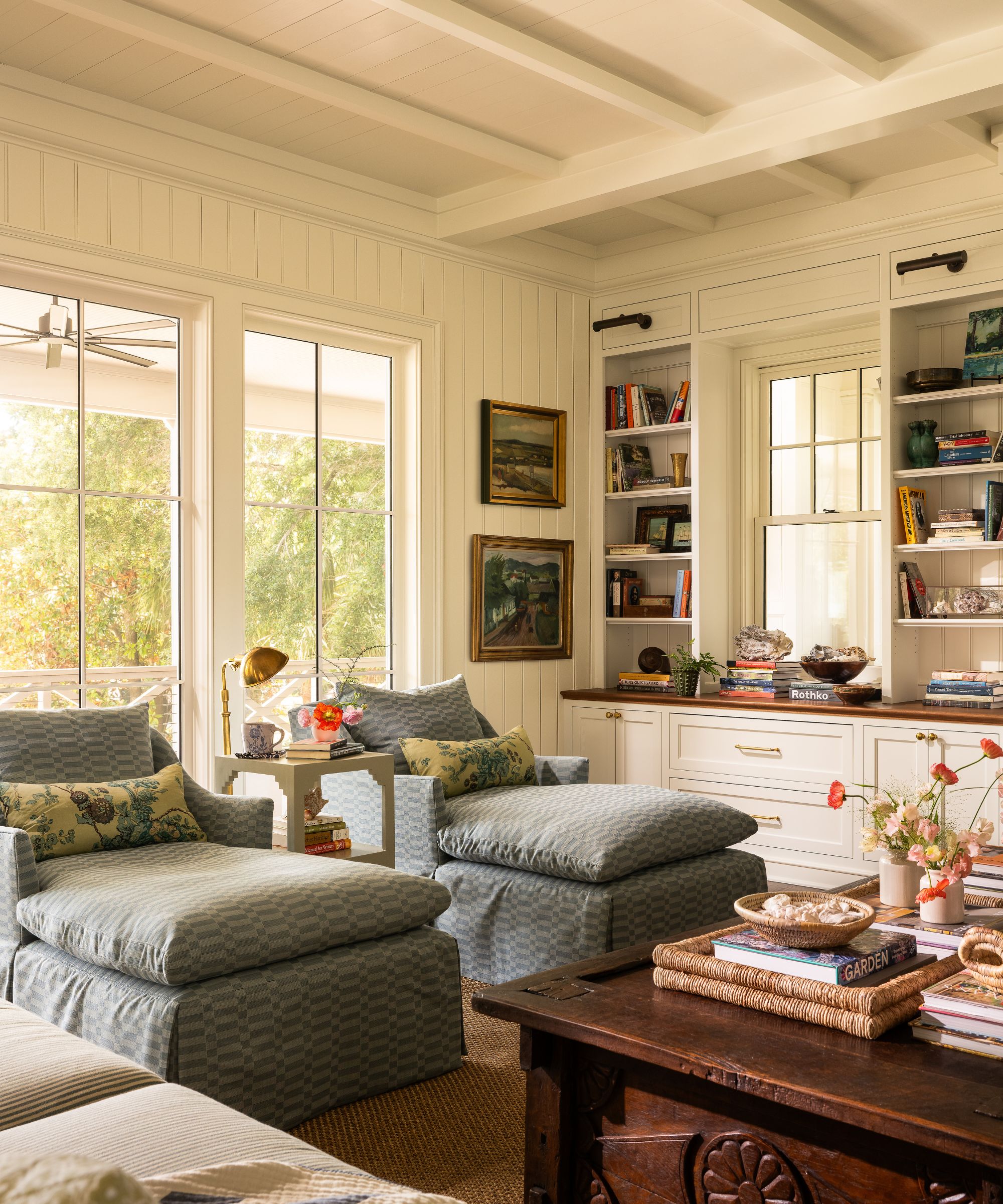 Coastal living room with ship lap white walls and beams, pale blue chaise lounges, mahogany coffee table and built-in bookcases with vintage books and trinkets