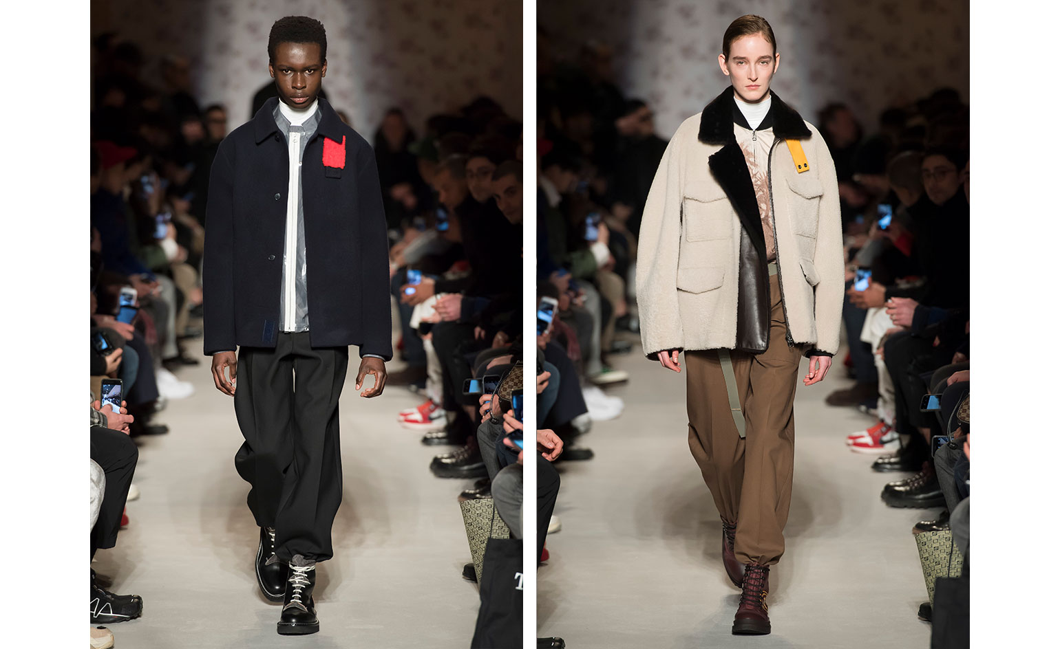 Catwalk view of two models wearing looks from OAMC's collection. One model is wearing a grey and white piece, blue jacket with red element, black trousers and black boots. And the second model is wearing a light coloured piece with darker coloured design and black collar, a cream jacket with black fur and yellow element, brown trousers, green belt and dark red boots. There are people seated on both sides of the catwalk